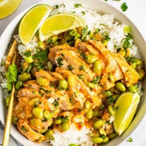 A bowl of white rice topped with sliced chicken, soybeans, and a creamy sauce, garnished with lime wedges and cilantro. A pair of gold chopsticks rests beside the bowl. Lime slices are scattered around.