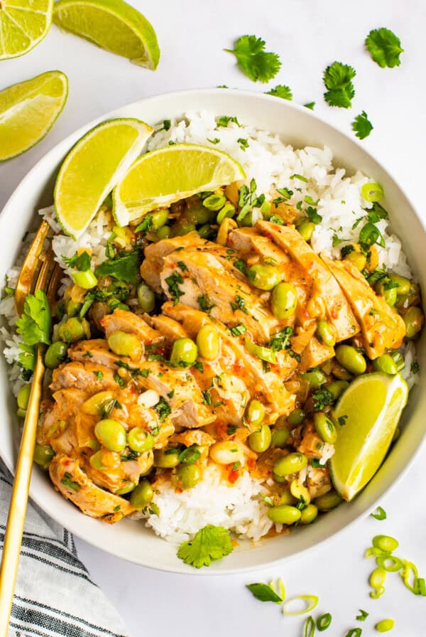 A bowl of white rice topped with sliced chicken, soybeans, and a creamy sauce, garnished with lime wedges and cilantro. A pair of gold chopsticks rests beside the bowl. Lime slices are scattered around.