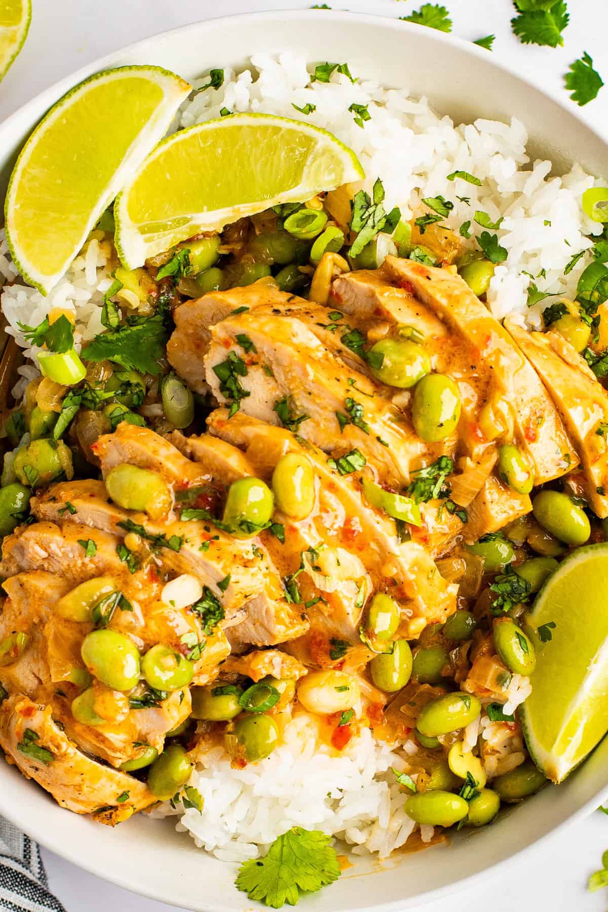 A bowl of white rice topped with sliced cooked chicken, edamame, and a creamy sauce, garnished with lime wedges and cilantro.