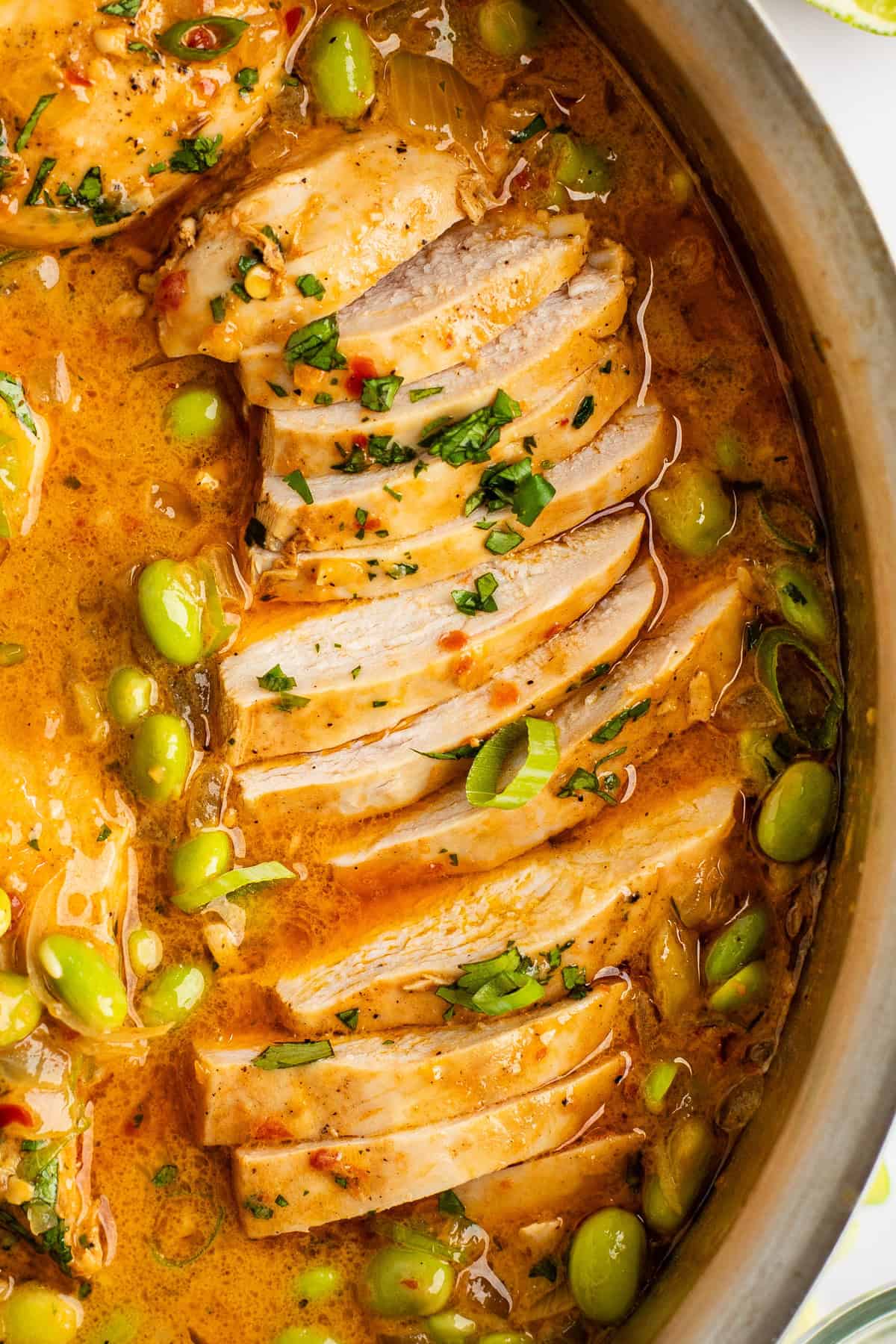 Close-up of sliced cooked chicken in a pan with a creamy, orange-colored sauce, garnished with chopped herbs and green peas.