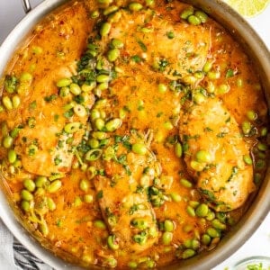 A stainless steel pan filled with cooked fish fillets in a vibrant orange sauce, garnished with chopped herbs and edamame beans. Lime wedges and a striped kitchen towel are placed beside the pan.