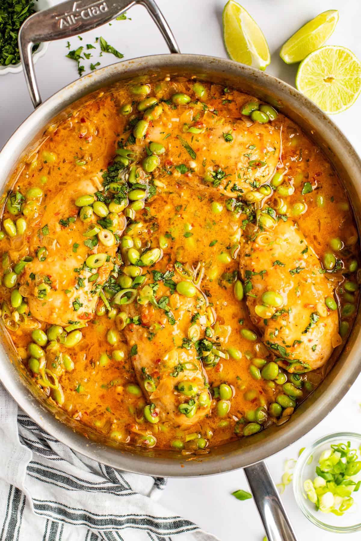 A stainless steel pan filled with cooked fish fillets in a vibrant orange sauce, garnished with chopped herbs and edamame beans. Lime wedges and a striped kitchen towel are placed beside the pan.