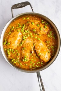 A stainless steel pan contains cooked chicken breasts in a golden sauce with green edamame.