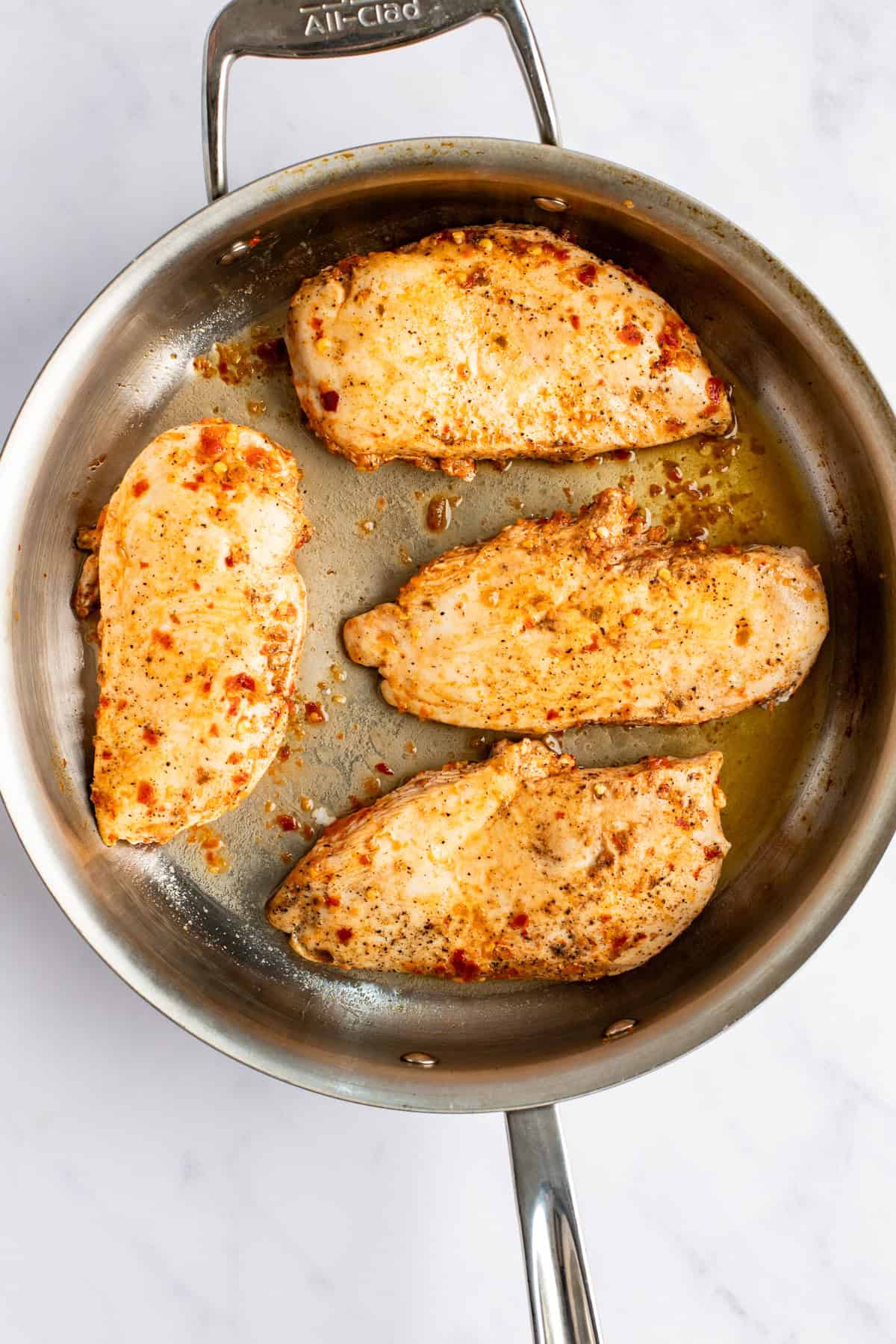 Four seasoned chicken breasts are cooking in a stainless steel skillet.