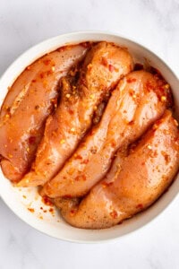 A bowl containing raw chicken breasts marinated with red pepper flakes and other spices.