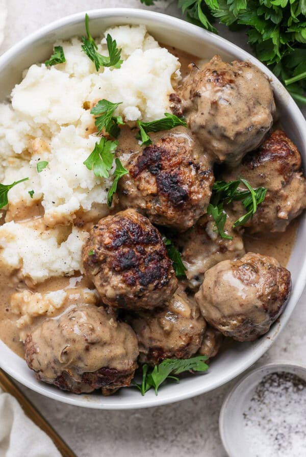 A bowl of meatballs in creamy sauce next to a serving of mashed potatoes. Fresh parsley garnishes the dish.