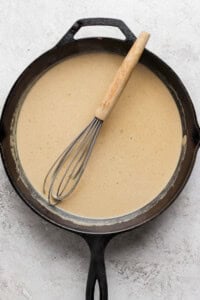 A cast iron skillet filled with a light brown sauce, with a whisk resting inside.