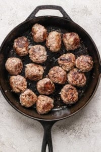 A cast-iron skillet contains 16 evenly browned meatballs on a textured light-colored surface.