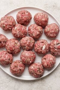 A plate filled with raw, evenly-sized meatballs, arranged in a circular pattern on a white surface.