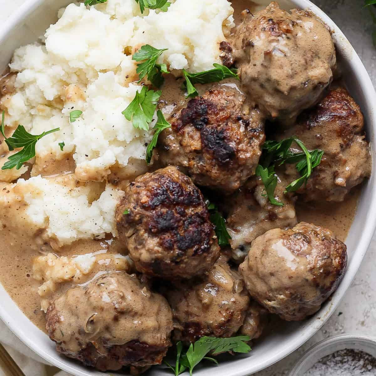 A bowl of meatballs in gravy, served with mashed potatoes, garnished with sprigs of fresh parsley.