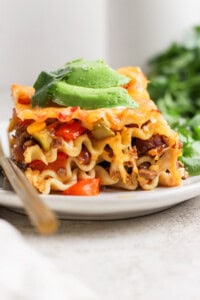 A close-up of a slice of lasagna topped with avocado slices on a white plate, with a fork beside it and green herbs in the background.