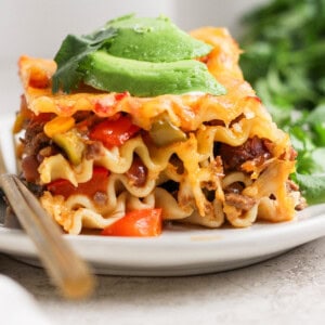 A close-up of a slice of lasagna topped with avocado slices on a white plate, with a fork beside it and green herbs in the background.