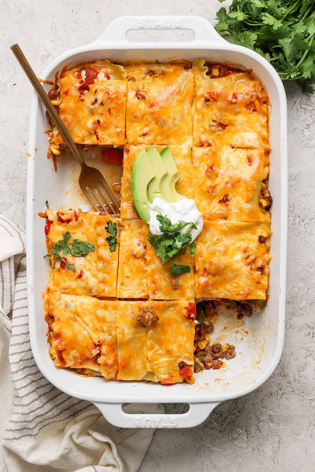 A baked casserole in a white rectangular dish is divided into squares, topped with melted cheese. Garnished with avocado slices, sour cream, and cilantro. A serving fork rests in one section.