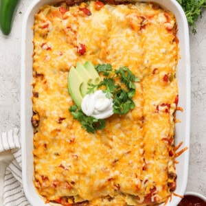 A baked casserole topped with melted cheese, avocado slices, sour cream, and cilantro, presented in a white baking dish. Fresh jalapeños, cilantro, and a dish of red sauce are beside it.