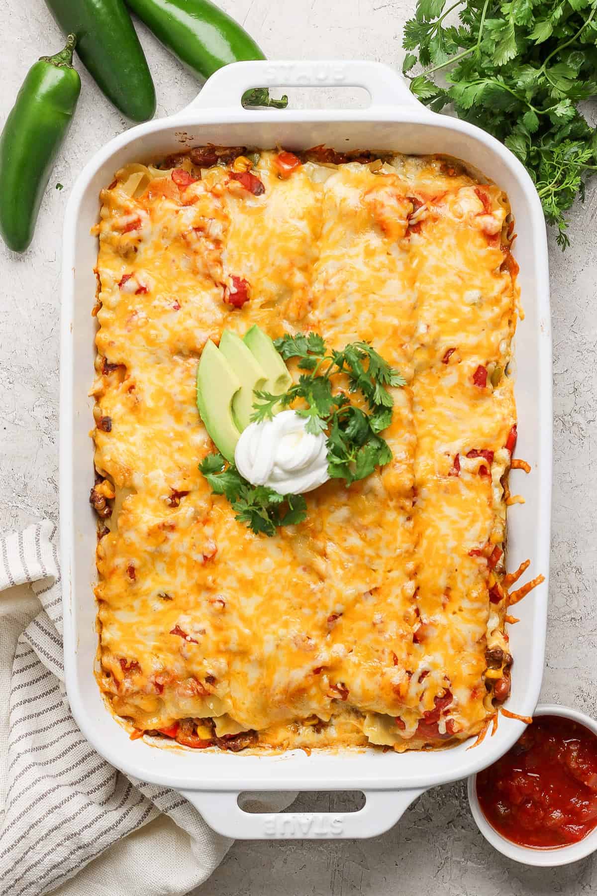 A baked casserole topped with melted cheese, avocado slices, sour cream, and cilantro, presented in a white baking dish. Fresh jalapeños, cilantro, and a dish of red sauce are beside it.