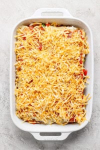 A white baking dish filled with an unbaked casserole topped with shredded cheese on a light-colored surface.