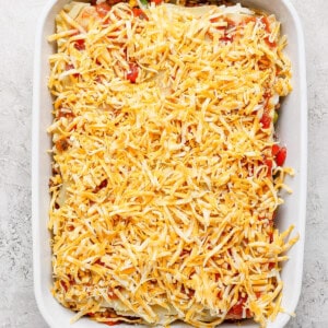 A white baking dish filled with an unbaked casserole topped with shredded cheese on a light-colored surface.