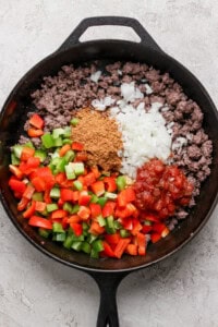 A cast iron skillet containing ground beef, diced green and red bell peppers, chopped onions, taco seasoning, and salsa.