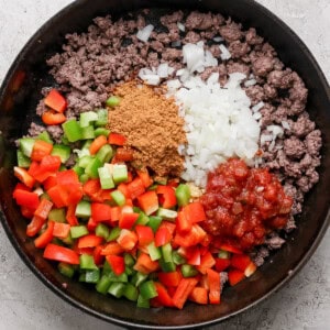 A cast iron skillet containing ground beef, diced green and red bell peppers, chopped onions, taco seasoning, and salsa.