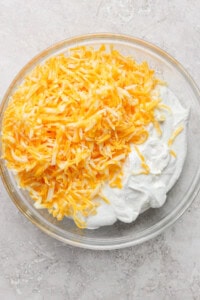 A clear glass bowl filled with shredded yellow and white cheese on top of a mound of white, creamy substance on a light-colored surface.