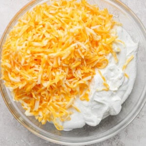 A clear glass bowl filled with shredded yellow and white cheese on top of a mound of white, creamy substance on a light-colored surface.