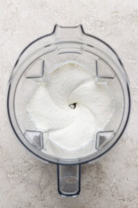 Top view of a clear blender jar containing a smooth, creamy white mixture, likely preparing a food or beverage, on a light textured surface.