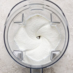 Top view of a clear blender jar containing a smooth, creamy white mixture, likely preparing a food or beverage, on a light textured surface.