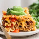 A slice of lasagna topped with avocado slices and garnished with cilantro, served on a white plate with a fork.