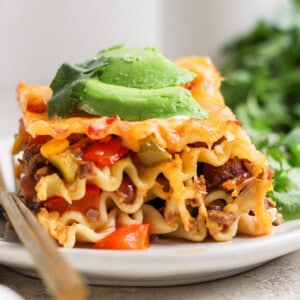 A slice of lasagna topped with avocado slices and garnished with cilantro, served on a white plate with a fork.