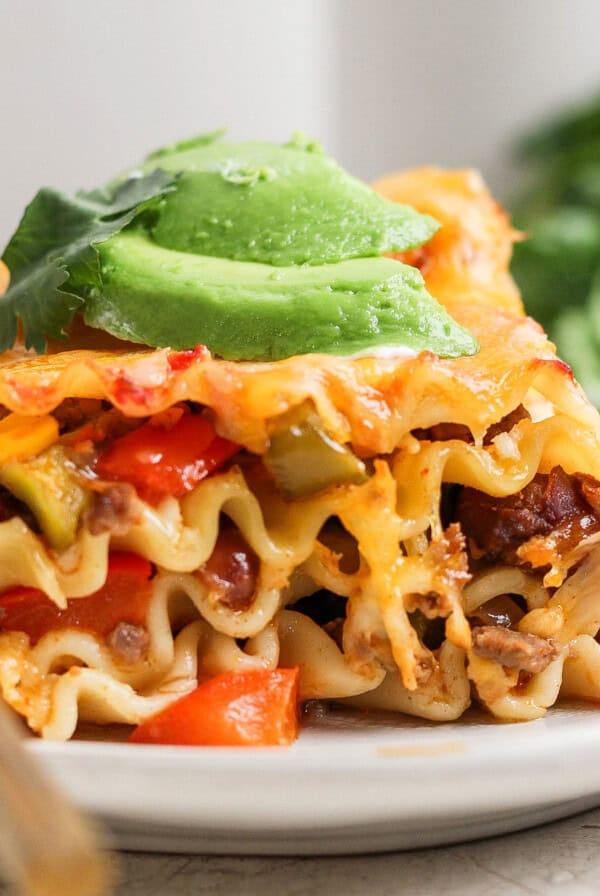 A slice of lasagna topped with avocado slices and garnished with cilantro, served on a white plate with a fork.