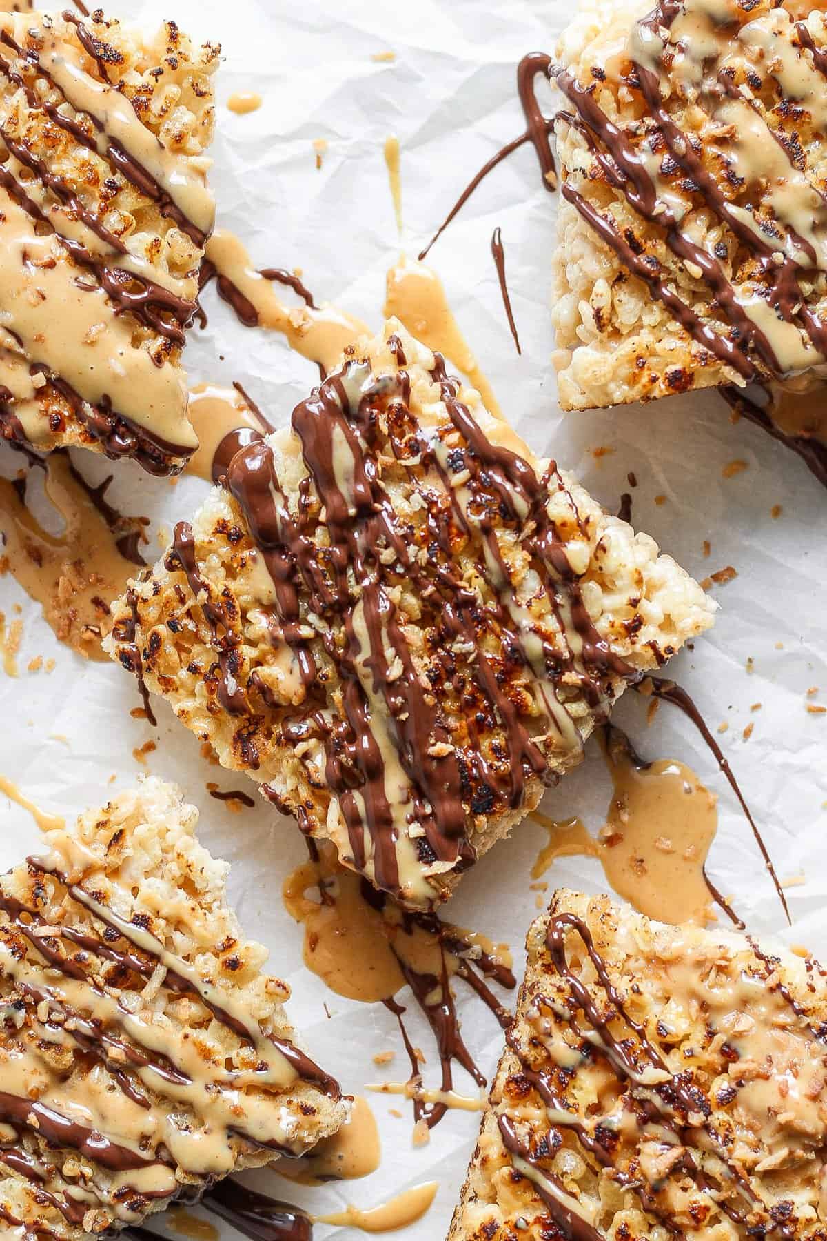 Close-up of five toasted Rice Krispie treats drizzled with chocolate and peanut butter, arranged on parchment paper. The treats are topped with crushed nuts.