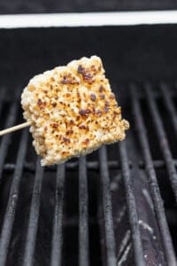 A square rice cake on a skewer is being grilled over open flames, showing light charring and crispness, reminiscent of toasted rice Krispie treats.