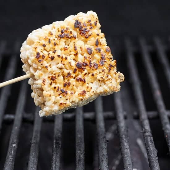 A square rice cake on a skewer is being grilled over open flames, showing light charring and crispness, reminiscent of toasted rice Krispie treats.