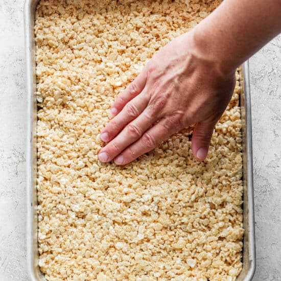 A hand pressing down on a tray filled with warm, toasted Rice Krispie treats mixture.