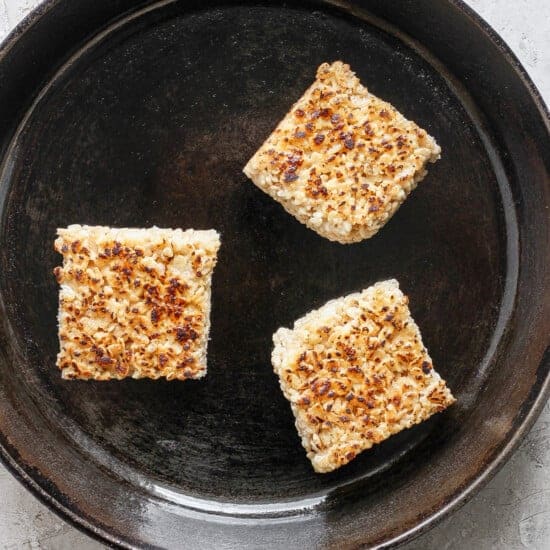 Three toasted Rice Krispie treats in a cast iron skillet on a light textured surface.