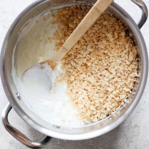 A pot filled with melted marshmallows and some toasted Rice Krispie treats, with a wooden spoon resting inside, on a light-colored surface.