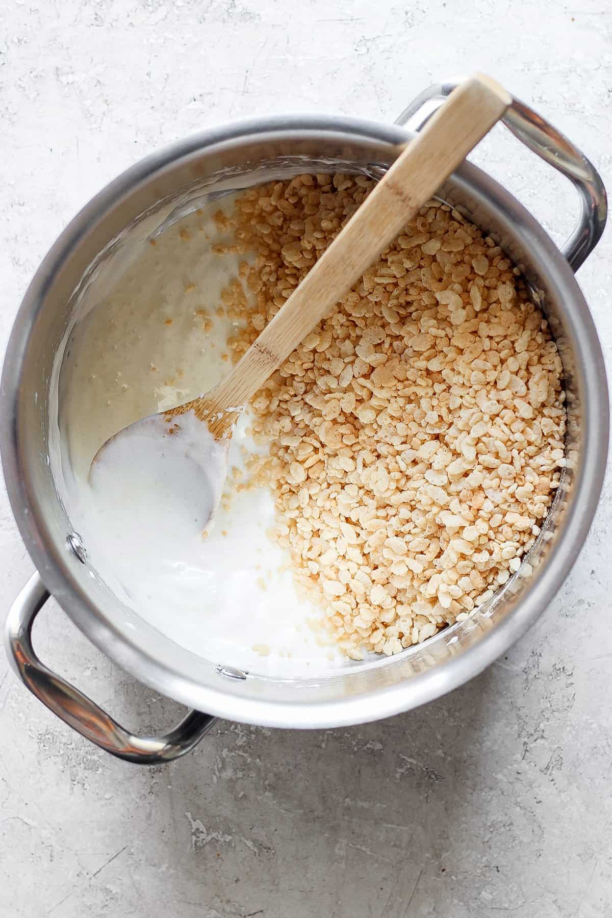 A pot filled with melted marshmallows and some toasted Rice Krispie treats, with a wooden spoon resting inside, on a light-colored surface.
