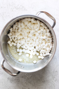 A metal pot containing partially melted mini marshmallows on a light-colored surface, ready to be transformed into delicious toasted Rice Krispie treats.