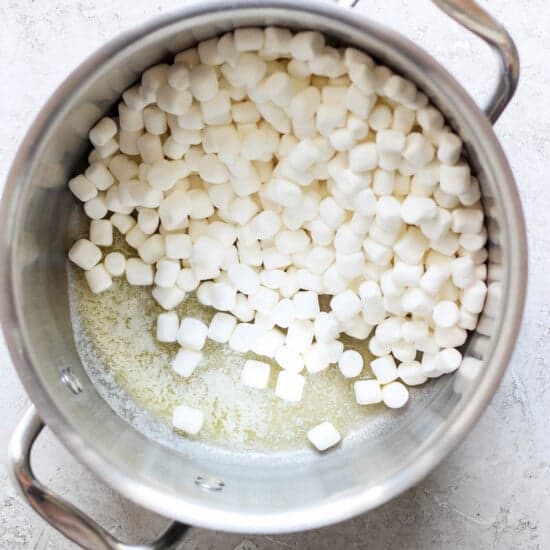 A metal pot containing partially melted mini marshmallows on a light-colored surface, ready to be transformed into delicious toasted Rice Krispie treats.
