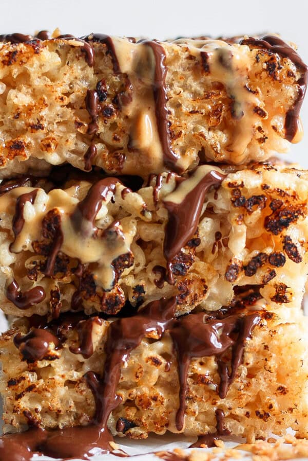 Stacked toasted rice krispie treats drizzled with chocolate and caramel sauces on a white surface.