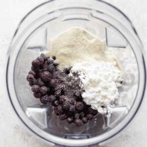 A blender contains frozen blueberries, cottage cheese, protein powder, and chia seeds, seen from a top view.
