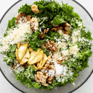 A bowl containing kale, walnuts, cheese shreds, and artichokes, prepared for a salad.