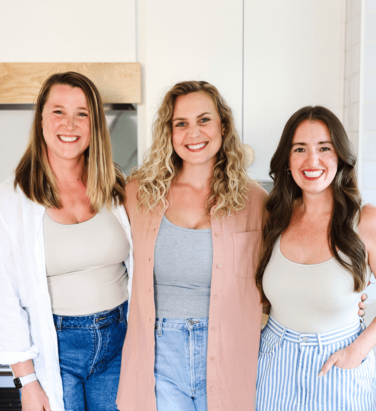 Three women, embodying the spirit of Fit Foodie Finds, smile while standing close together in casual attire for an indoor photo. One wears jeans and a white shirt, another a pink shirt, and the third sports stylish striped pants.