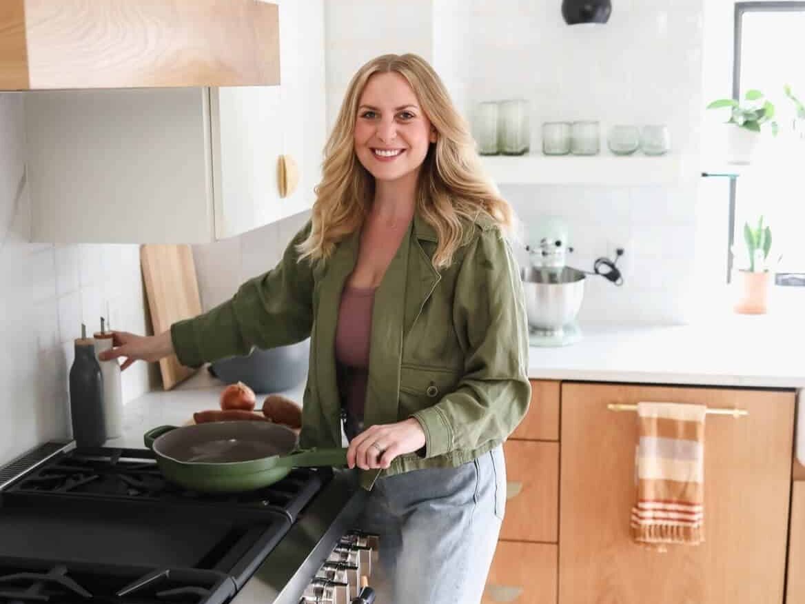 A woman stands in a kitchen, holding a frying pan on the stove with one hand and an oil bottle with the other. Natural light illuminates the modern kitchen featuring wooden cabinets and white countertops. It's all about fit foodie finds in this beautifully lit space.