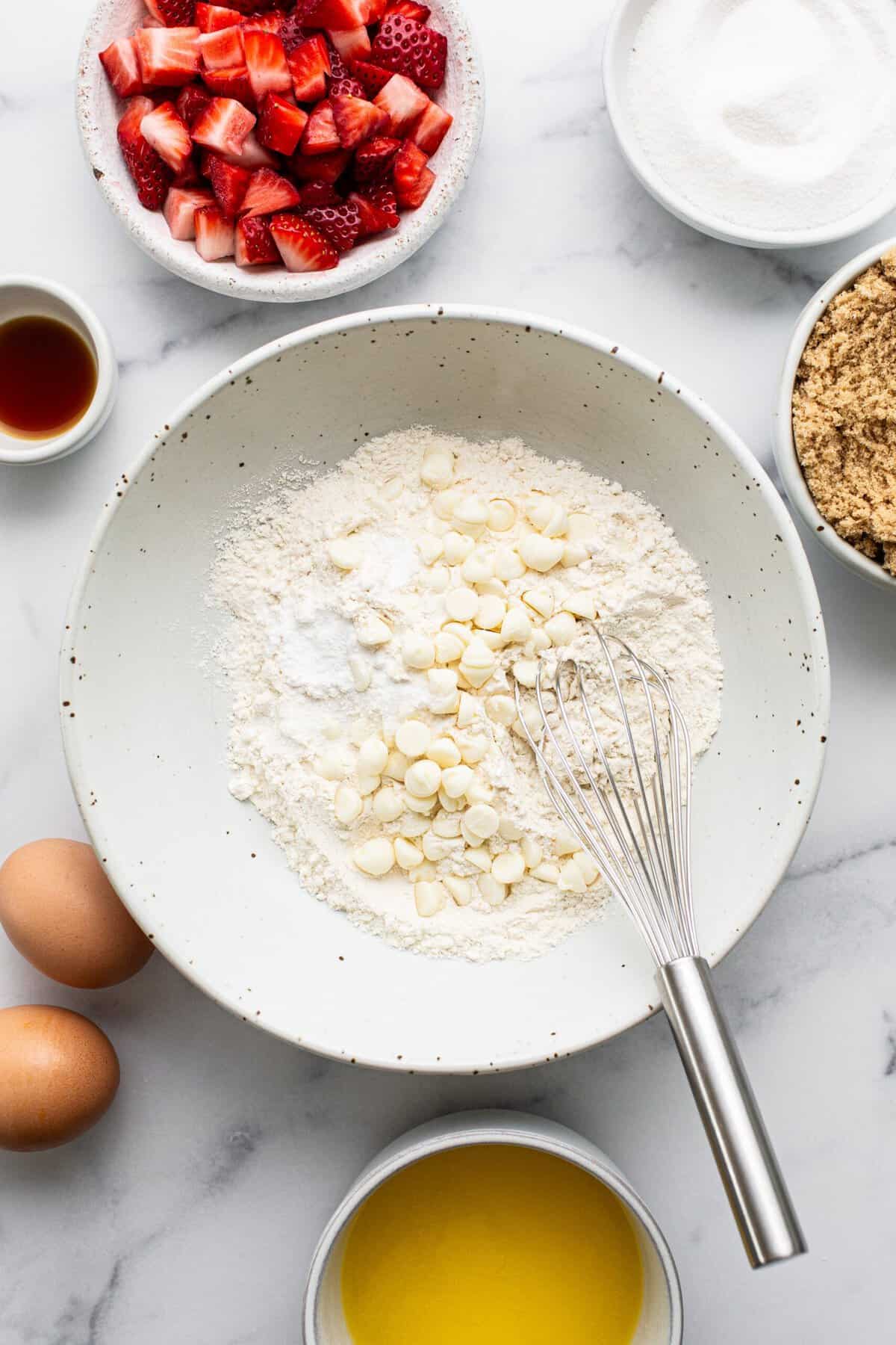 Top-down view of baking ingredients on a marble surface, perfect for making strawberry blondies, including a bowl of flour and white chocolate chips with a whisk, two eggs, strawberries, sugar, brown sugar, melted butter, and vanilla extract.