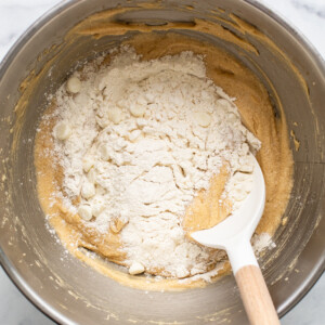 A mixing bowl containing partially mixed cookie dough with flour and white chocolate chips, resembling the beginnings of delectable strawberry blondies, and a white spatula resting inside.