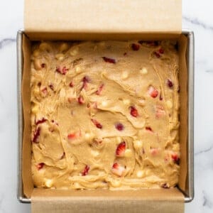 Square baking pan lined with parchment paper and filled with raw batter containing chunks of fruit, promising delicious strawberry blondies on a marble countertop.