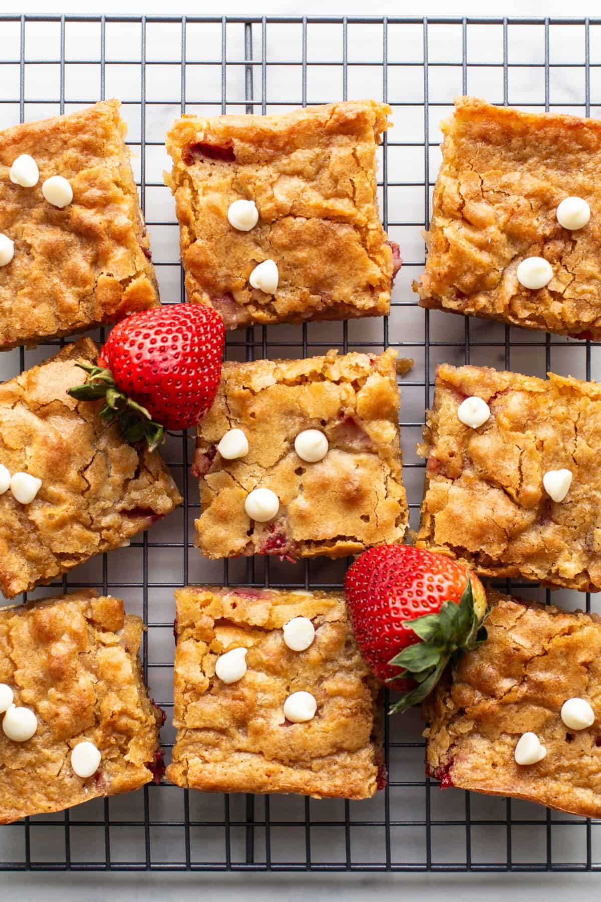 Nine square pieces of strawberry blondies with white chocolate chips and fresh strawberries rest on a cooling rack.