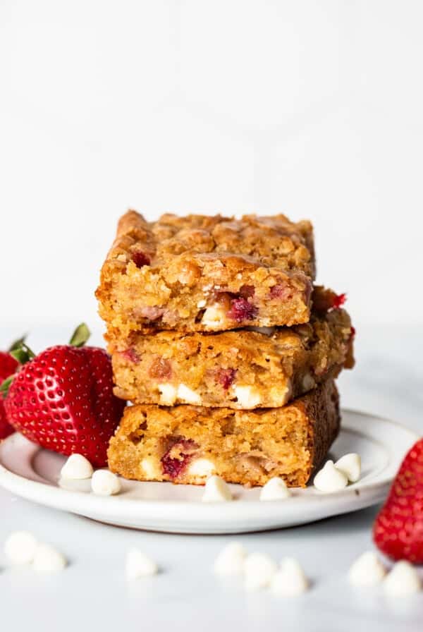 A plate of three stacked strawberry blondies containing white chocolate chips and strawberry pieces, surrounded by strawberries and white chocolate chips.