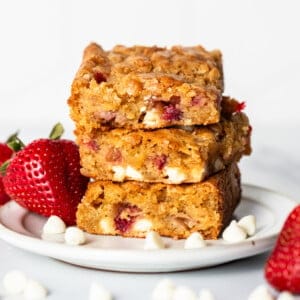 Three strawberry blondies, layered with white chocolate, are stacked on a white plate, surrounded by fresh strawberries and white chocolate chips.